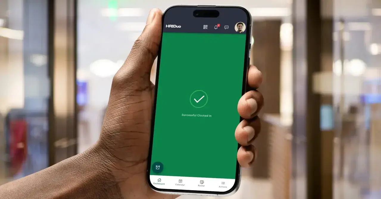 A hand holding a phone showing a successful clock-in on the HR:Duo app