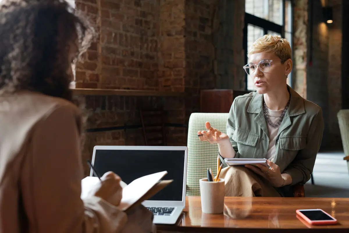 Two women taking notes on either side of a table during a formal workplace meeting