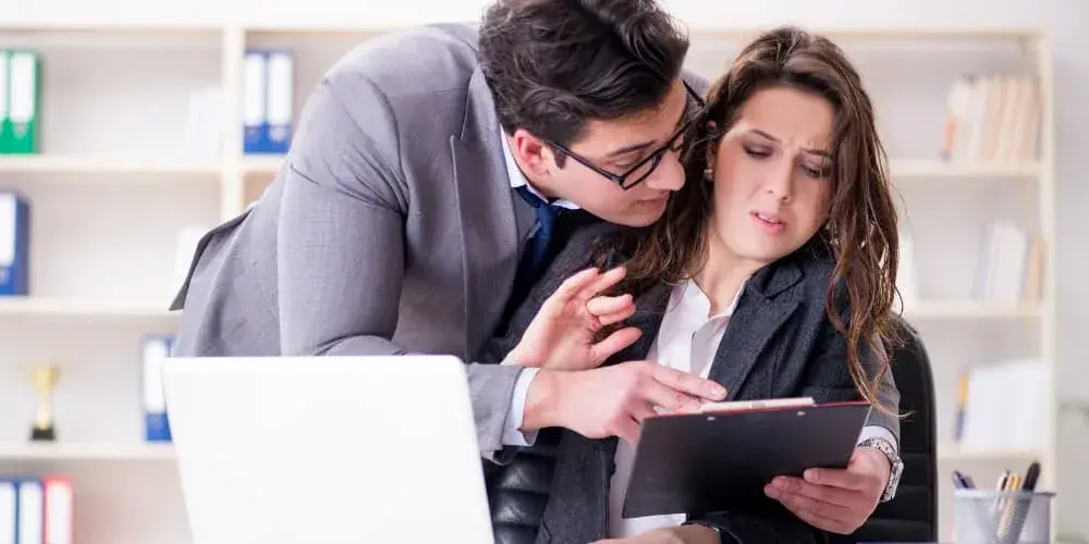 A man leans uncomfortably close to an uneasy colleague at her office desk